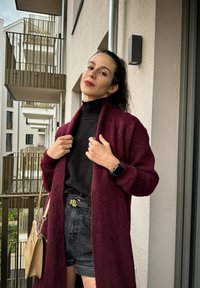 Μακρύ μπορντό κασκόλ από υφαντό πλέγμα, στρωματωμένο πάνω από ένα μαύρο ζιβάγκο, συνδυασμένο με μαύρες σορτς και μπεζ τσάντα.