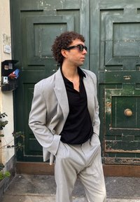 Muž v sivom obleku a čiernej košeli s okuliarmi na slnko stojí s rukami v kapsách pred opotrebovanými zelenými drevenými dverami.