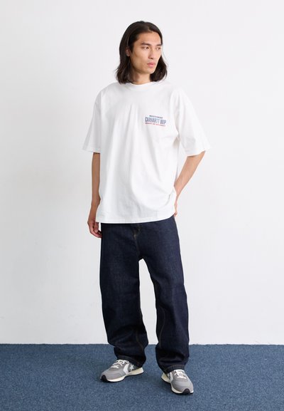 Camiseta de algodón blanca con un gráfico en púrpura y azul, combinada con pantalones de mezclilla azul oscuro. El modelo lleva zapatillas grises con detalles en negro.