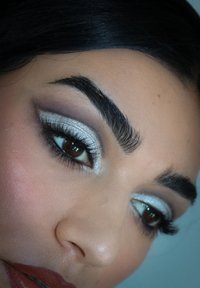 Nærbilde av et øye med sølvfarget øyenskygge, mørke vipper og definerte bryn. Myk hudtekstur og naturlig leppefarge er synlig.