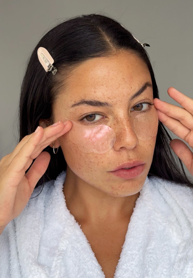Mujer de cabello oscuro con bata blanca aplica parches de gel para debajo de los ojos mientras sujeta el cabello con pinzas sobre un fondo neutro.