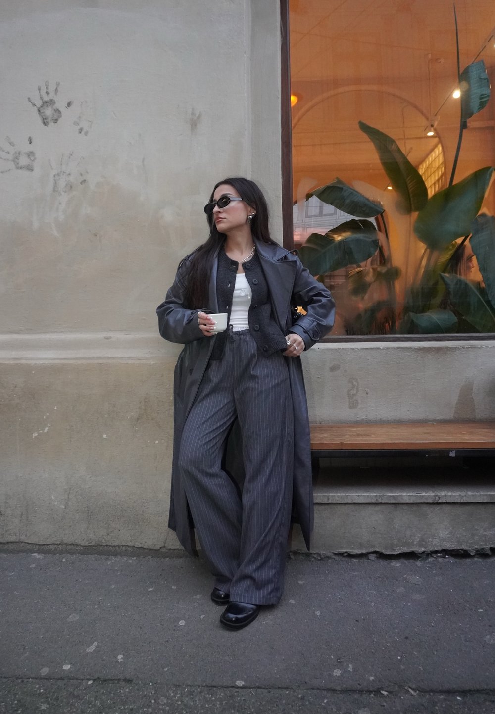 Longue veste trench-coat en cuir noir portée sur un cardigan en maille gris, un haut blanc et un pantalon large à fines rayures. Lunettes de soleil noires et tenant une tasse.