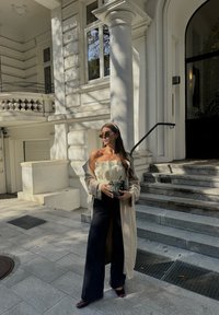 Μπεζ παλτό πάνω από κρεμ τοπ χωρίς τιράντες, φαρδιές μαύρες τζιν κι ένα σκούρο κόκκινο τακούνι. Κρατά μια μικρή τσάντα με σχέδιο. Σκαλοπάτια και κτίριο στο βάθος.