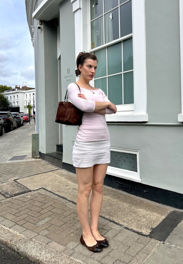 Mujer con top rosa claro y falda blanca está de pie con los brazos cruzados en la acera frente a un edificio gris con una gran ventana.