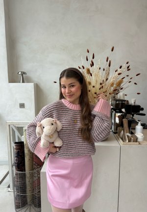 Mujer joven con falda rosa y suéter a rayas sosteniendo un elefante de peluche, de pie cerca de plantas secas y una cafetera en una habitación minimalista.