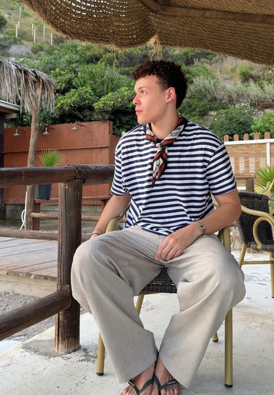 Camiseta a rayas en azul marino y blanco, pantalones de lino color beige claro, chanclas negras y una bufanda roja con estampado, sentado al aire libre en una silla de madera.