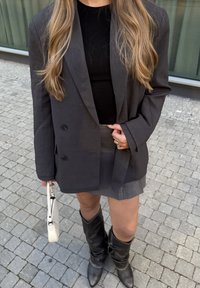 Fekete szabott blézer, testhezálló fekete felső, szürke réteges szoknya, térdfeletti fekete csizma hegyes orral, és egy kis fehér kézitáska.