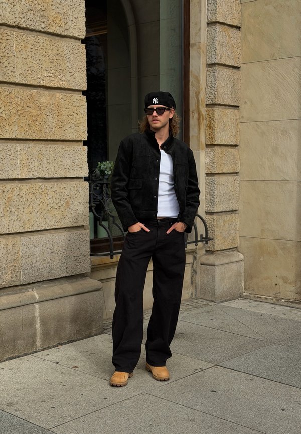 Un hombre con una chaqueta negra, pantalones de pernera ancha, gafas de sol y una gorra de los Yankees está de pie frente a un edificio de piedra con las manos en los bolsillos.