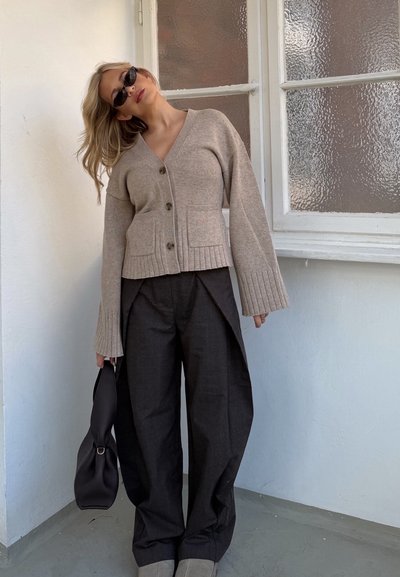 Mujer con cárdigan beige con botones, pantalones negros de pierna ancha, gafas de sol, sosteniendo bolsa negra, de pie en el interior cerca de una ventana de vidrio texturizado.