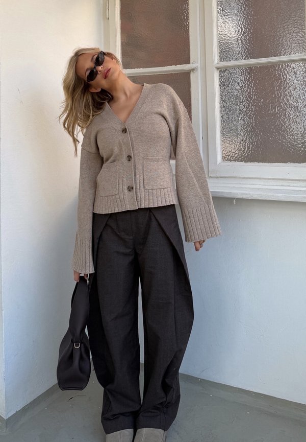 Nő bézs gombos kardigánban, széles szárú fekete nadrágban, napszemüvegben, fekete táskát tartva, beltéren, egy mintás üvegablak mellett áll.