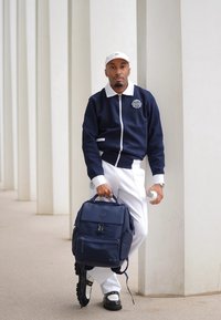 Giacca a zip blu navy con colletto bianco, pantaloni bianchi e scarpe nere. Tiene uno zaino blu navy e una bottiglia d'acqua, in piedi vicino a colonne bianche.