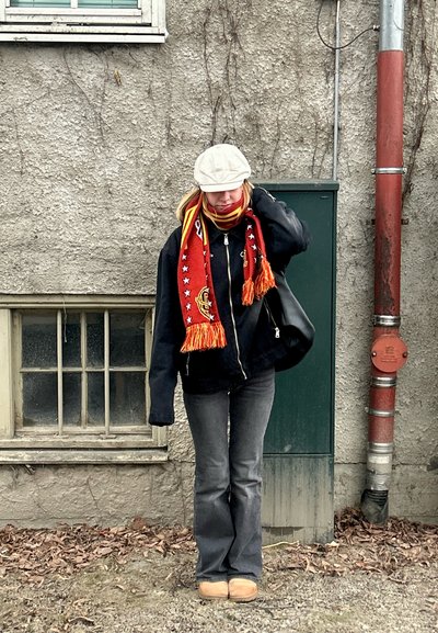 Persona con chaqueta negra, jeans grises y una bufanda roja con patrones amarillos de estrellas. Un gorro beige y un bolso negro completan el estilo.