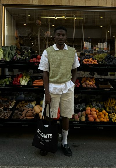 Chaleco de punto verde sobre una camisa blanca, pantalones cortos blancos, zapatos negros y bolsa tote negra. Frutas y verduras frescas en el fondo.