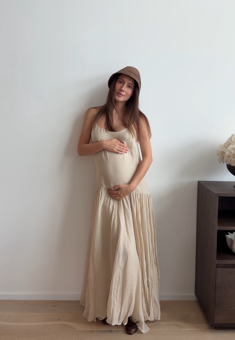 Femme enceinte en robe beige et chapeau marron tenant doucement son ventre, debout contre un mur blanc à côté d'une armoire en bois foncé avec des fleurs.