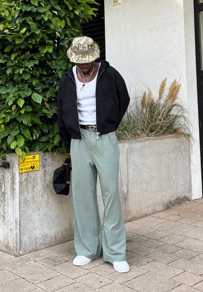 Sudadera negra con cremallera, camiseta de tirantes blanca, pantalones anchos verdes, zapatillas blancas, sombrero tipo cubo de camuflaje, sosteniendo una bolsa negra, de pie junto a un follaje.