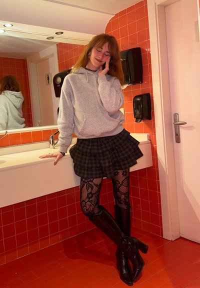 Joven con cabello rojo apoyado en el lavabo de un baño con azulejos rojos, vistiendo una sudadera con capucha gris, falda de cuadros negra, medias de encaje y botas negras.