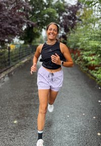 Kvinne som smiler og løper på en våt asfaltsti omkranset av trær med grønne og lilla blader, iført svart tank topp, lyse shorts og hvite joggesko.