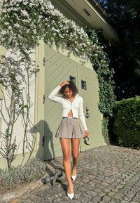 Cardigan court à côtes blanches avec fermeture éclair à l'avant, jupe patineuse beige avec cordon de serrage, et escarpins blancs à bout pointu sur un chemin en pavés.