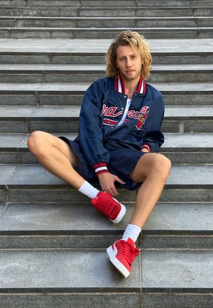 Chaqueta bomber de la marina con detalles en rojo y blanco, pantalones cortos de mezclilla y zapatillas rojas con suelas blancas, sentada en unos escalones de piedra.