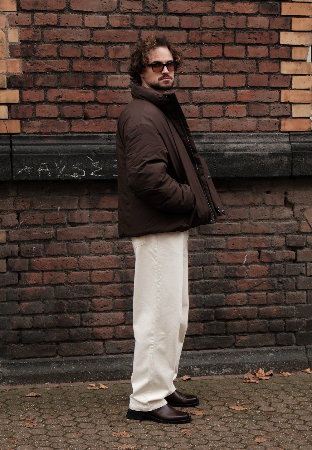Brown puffer jacket, cream wide-leg trousers, and brown leather boots. Standing against a brick wall, with a textured fabric and casual fit.