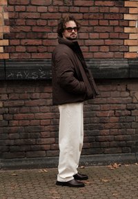 Brown puffer jacket, cream wide-leg trousers, and brown leather boots. Standing against a brick wall, with a textured fabric and casual fit.