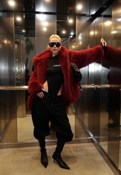 Mujer con gafas de sol negras, body negro, abrigo de piel rojo, pantalones negros y botas de tacón posando con un brazo extendido dentro de un ascensor con espejos.