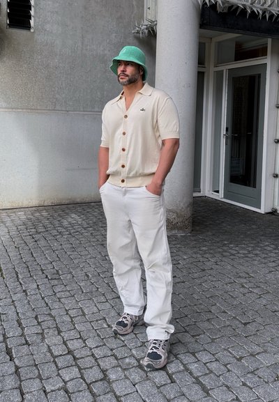 Camisa beige de mangas cortas con botones marrones, logo bordado, pantalones blancos, zapatillas grises y un sombrero de cubo verde, de pie sobre adoquines.