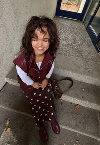 Vīnogu krāsas mākslīgās ādas veste, balta krekla ar garām piedurknēm, brūna punktainā svārki, tumši sarkani zābaki un rakstaina soma uz akmens kāpnēm.