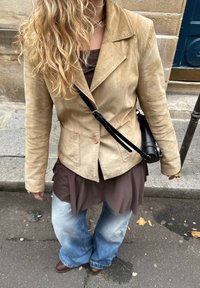Blonde persoon in een tan suede jacket, bruine top, wijde vervaagde jeans en bruine schoenen, staand op een stoep in de stad.