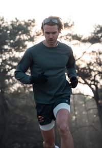 T-shirt de course à manches longues vert foncé avec surface texturée, associé à un short à motifs en noir et blanc avec des accents orange. Gants noirs portés.