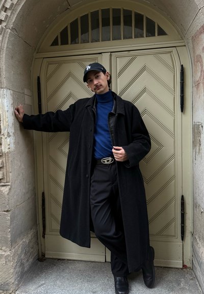 Largo abrigo negro sobre un cuello de tortuga azul, pantalones negros y una gorra. Hebilla de cinturón plateada y zapatos negros. De pie junto a una puerta texturizada.