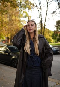 Brązowy skórzany płaszcz trenczowy noszony na dżinsowy strój, z paskiem w talii. W tle widać zamazane drzewa i zaparkowane samochody.