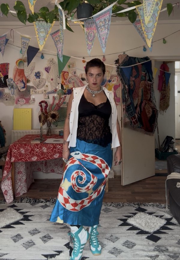 Une personne pose dans une pièce décorée, vêtue d’un haut noir en dentelle, d’un gilet blanc, d’une jupe bleue à motifs et de chaussures montantes turquoise.