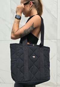 Μαύρη τακτοποιημένη τσάντα με επένδυση από μαύρο ύφασμα, λεπτομέρεια λογότυπου, ενισχυμένες λαβές και εξωτερική τσέπη. Ο σχεδιασμός διαθέτει ορθογώνιο σχήμα.