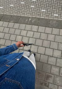 Occhiali da sole neri tenuti in una mano, giacca di jeans, pantaloni della tuta grigi, in piedi su un pavimento in mattoni grigi testurizzati con un motivo a pesce spada.