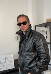 Veste en cuir noire avec un col montant, une fermeture éclair et des poches avant. Le mannequin porte des lunettes de soleil foncées, avec une texture visible sur la veste.