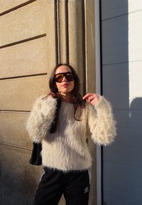 Luddig, krämfärgad tröja med långa ärmar, kombinerad med svarta mjukisbyxor med vita ränder och solglasögon. Står mot en texturerad vägg.