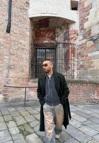 Homme portant des lunettes de soleil, des écouteurs, un manteau sombre et un pull gris, marchant sur un pavé en pierre près d'un mur en briques et pierres usées avec une fenêtre grillagée.