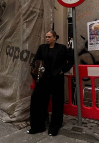 Black oversized blazer with wide-leg trousers, complemented by a brown bag. Silver accents and a bun hairstyle, set against a construction backdrop.