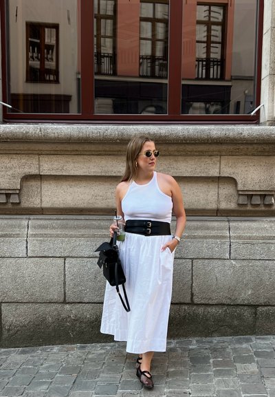 Vestido blanco sin mangas con un cinturón negro que tiene dos hebillas, combinado con zapatos negros y un bolso negro; sosteniendo una bebida. Fondo de pared de piedra.