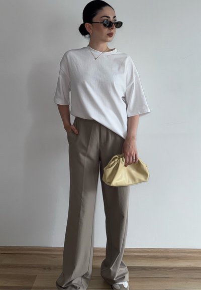 Mujer con el pelo recogido en un moño, lleva gafas de sol negras, camiseta blanca oversize, pantalones beige holgados, sosteniendo un pequeño bolso amarillo contra una pared lisa.