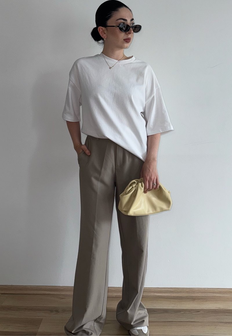Femme aux cheveux en chignon, portant des lunettes de soleil noires, un t-shirt blanc oversize, un pantalon beige ample, tenant un petit sac à main jaune contre un mur uni.