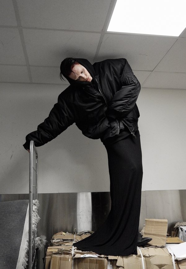 Oversized black puffer jacket with a high collar, paired with a fitted black skirt. Subject poses against a backdrop of cardboard boxes.