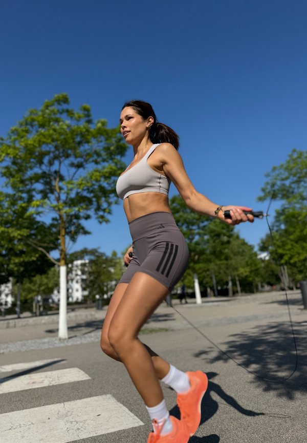 Femme portant des vêtements de sport et des chaussures orange vif sautant à la corde à l'extérieur sur une surface pavée avec des arbres et un ciel bleu clair.