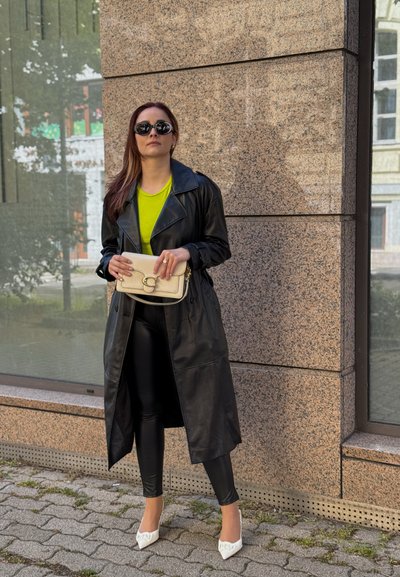 Abrigo de cuero negro sobre una blusa verde brillante, combinado con leggings negros brillantes y tacones blancos de punta. Sosteniendo un bolso clutch beige.