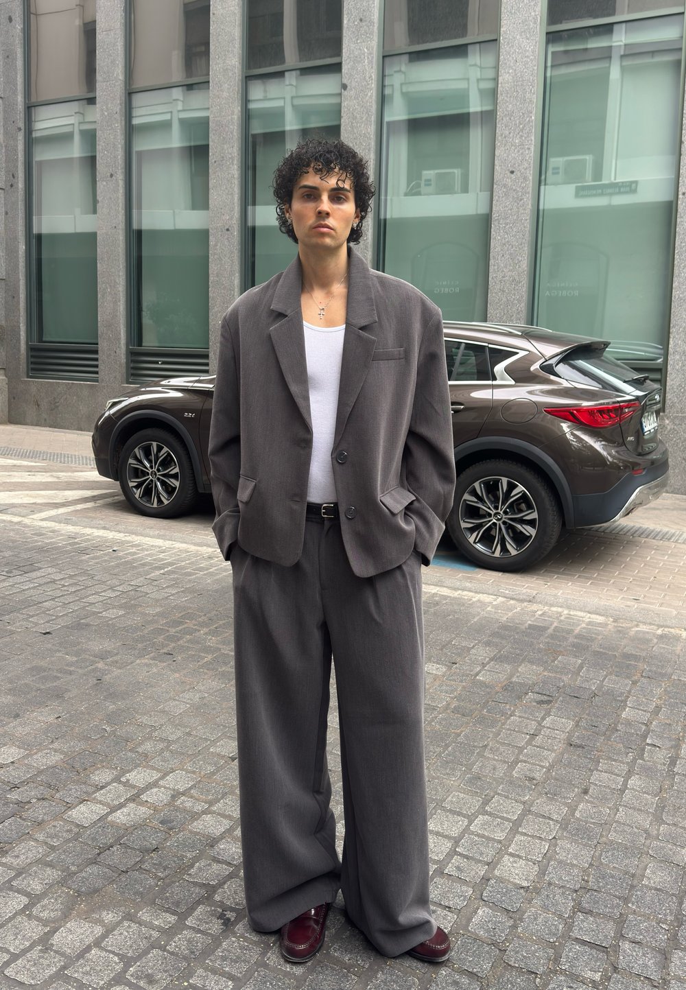Bărbat cu păr creț, purtând un costum gri supradimensionat, maiou alb și pantofi bordo, stă pe o stradă pietruită, cu un SUV maro și o clădire modernă în spate.
