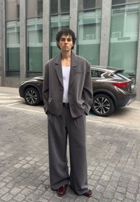 Bărbat cu păr creț, purtând un costum gri supradimensionat, maiou alb și pantofi bordo, stă pe o stradă pietruită, cu un SUV maro și o clădire modernă în spate.