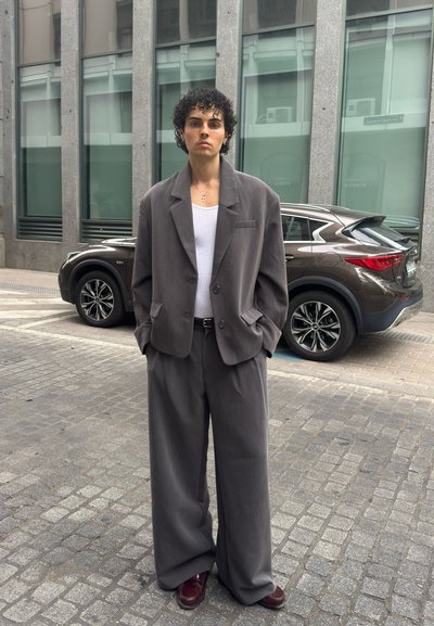 Hombre con cabello rizado que lleva un traje gris de gran tamaño, camiseta sin mangas blanca y zapatos granate, de pie en una calle empedrada con un SUV marrón y un edificio moderno detrás.