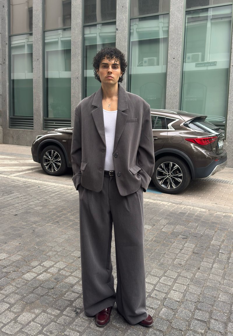 Homme aux cheveux bouclés portant un costume gris oversized, un débardeur blanc et des chaussures bordeaux, debout sur une rue pavée avec un SUV marron et un bâtiment moderne en arrière-plan.