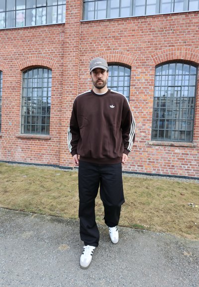 Hombre con sudadera Adidas marrón oscuro, pantalones negros, zapatillas blancas y gorra gris caminando por un camino frente a un edificio de ladrillos con grandes ventanas.